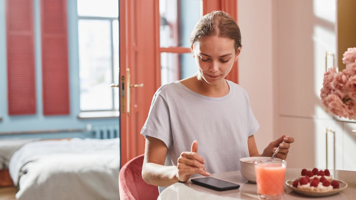 Young woman enjoying breakfast at home while using her smartphone. Bright, sunny morning ambiance.